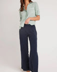 A woman stands against a plain light background wearing a Bella Dahl light green button-up shirt with rolled sleeves, navy wide-leg denim-look pants, and tan flat sandals. Her hair is loose and wavy, with her face partially out of frame.