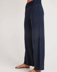 A person in profile wears Bella Dahl’s navy blue wide-leg pants in denim look, featuring a smocked waistband. Their feet are in flat tan sandals with gold crisscross straps. The plain light background highlights only their lower torso and legs.