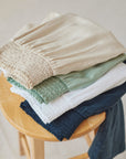 A neatly folded stack of five Bella Dahl weite Hosen in beige, light green, white, light blue, and navy sits on a wooden stool, showing smocked waistbands—a soft display of Lyocell and Denimoptik styles.