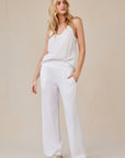 A woman with long blonde hair stands against a beige background, wearing a loose white Bella Dahl camisole and Lyocell trousers. With her hands in her pockets, tan slide sandals, and a relaxed pose, she exudes effortless style.