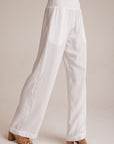 A person stands against a light background wearing loose-fitting, white, wide-leg Bella Dahl Lyocell pants with a smocked elastic waistband. The slightly sheer pants are paired with tan, open-toed block-heeled sandals. Upper body is not visible.