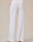 A person faces away, displaying the back of loose-fitting white Bella Dahl Lyocell wide-leg pants with a broad smocked waistband in denim look. They wear tan slide sandals against a light beige neutral backdrop; both floor and pants are light-colored.