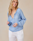 A smiling woman with long blonde hair models a light blue Bella Dahl Lyocell blouse with tie strings and a deep V-neck, paired with white pants. She stands against a plain beige background, looking slightly to the side.