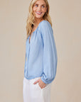 A woman with long blonde hair smiles, looking over her shoulder. She wears a light blue Bella Dahl Lyocell blouse with balloon sleeves and white pants, one hand in her pocket, standing against a soft beige background.