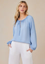 A woman with long blonde hair wears a light blue Bella Dahl Lyocell blouse with balloon sleeves and a tie neckline, paired with relaxed white pants. She stands with hands in her pockets, gazing to the side against a plain beige background.