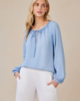 A woman with long blonde hair wears a light blue Bella Dahl Lyocell blouse with balloon sleeves and a tie neckline, paired with relaxed white pants. She stands with hands in her pockets, gazing to the side against a plain beige background.