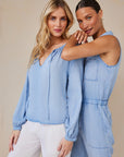 Two women pose against a neutral background, both in light blue Bella Dahl Lyocell denim tops—one wears a balloon-sleeve blouse with white pants, the other a sleeveless jumpsuit. Smiling softly, one rests her arm on the others shoulder.