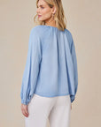 A woman with loose blonde hair stands against a beige background, wearing a light blue Bella Dahl Lyocell blouse with balloon sleeves and gathered neckline and cuffs, paired with white pants. She faces away, her head turned slightly to the side.