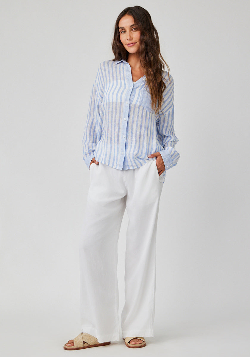 A woman with long wavy brown hair stands against a light background, wearing a Bella Dahl striped light blue-white linen-blend shirt, wide-leg white pants, and beige slide sandals. Her hands are in her pockets.