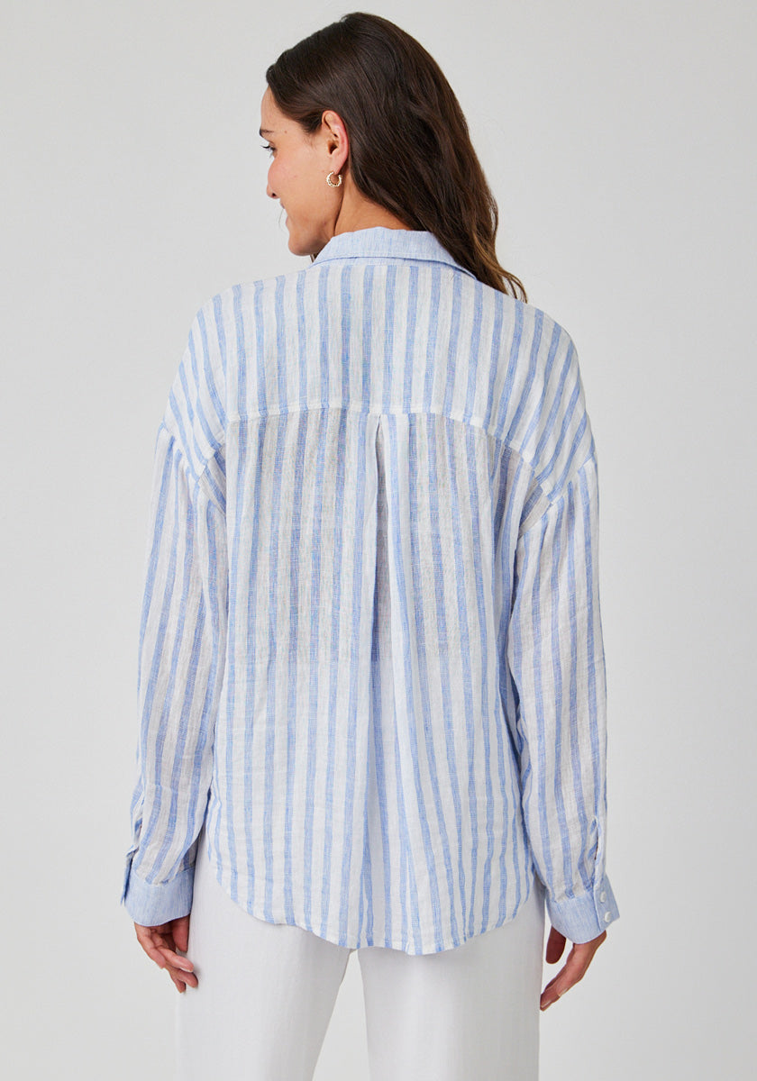 A woman faces away, wearing a Bella Dahl loose, long-sleeved linen-blend shirt with vertical blue and white stripes. Her brown hair is down, and she pairs the blouse with white pants against a simple light background.