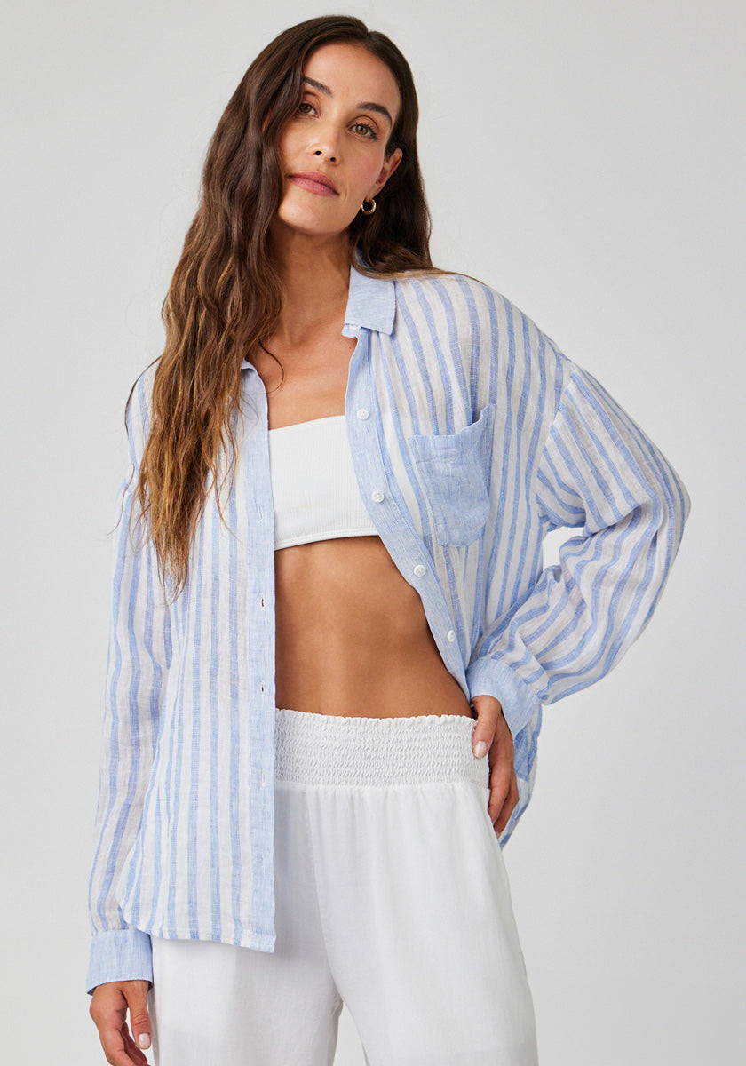 A woman with long wavy brown hair poses against a plain background, wearing a Bella Dahl gestreifte Bluse im Leinenmix über einem weißen Bandeau-Top und lockeren weißen Hosen. Sie wirkt entspannt und hat eine Hand in der Tasche.