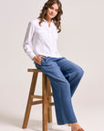 A woman with wavy brown hair sits on a wooden stool, wearing a white Lyocell Bluse in Jacken-Stil by Bella Dahl, wide-leg blue jeans, and brown sandals. She smiles slightly with her left hand in her pocket against a plain light gray background.