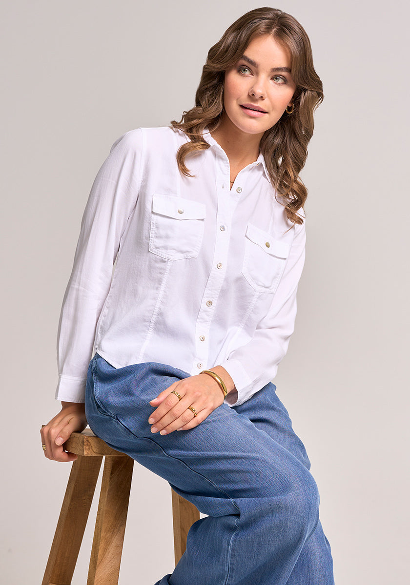 A woman with wavy brown hair sits on a wooden stool, wearing a white Bella Dahl Lyocell Bluse im Jacken-Stil with front pockets and loose blue jeans. She accessorizes with gold hoop earrings, a bracelet, and ring, gazing right with a relaxed expression.
