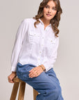 A woman with wavy brown hair sits on a wooden stool, wearing a white Bella Dahl Lyocell Bluse im Jacken-Stil with front pockets and loose blue jeans. She accessorizes with gold hoop earrings, a bracelet, and ring, gazing right with a relaxed expression.