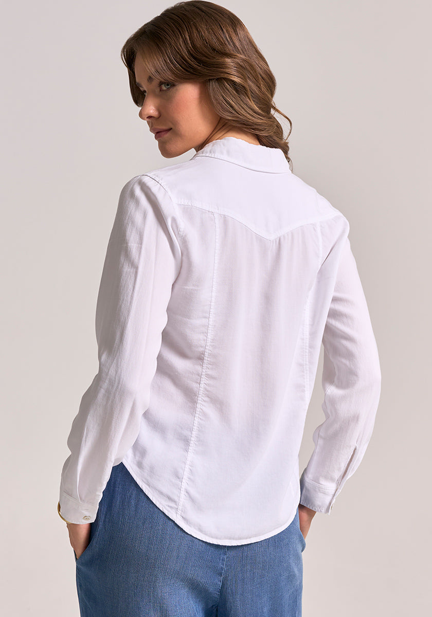 A woman with light brown, wavy hair looks over her shoulder. She wears a long-sleeved Bella Dahl Lyocell Bluse im Jacken-Stil and blue pants, hands in pockets, against a plain, light background.