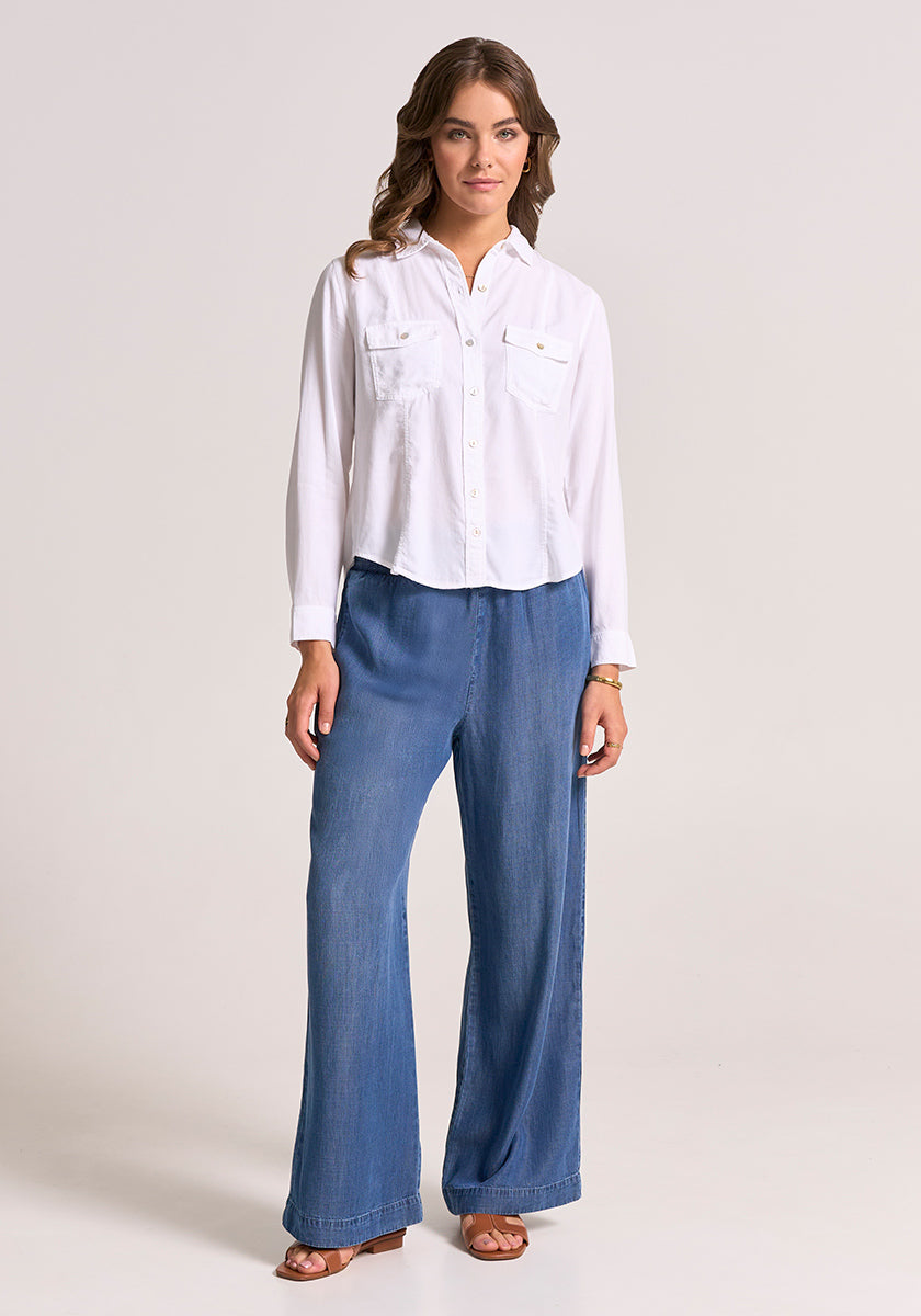 A woman with wavy brown hair and a neutral expression stands against a light background wearing a white Bella Dahl Lyocell blouse with front pockets and button placket, loose wide-leg blue jeans, and brown sandals.