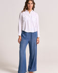 A woman with wavy brown hair and a neutral expression stands against a light background wearing a white Bella Dahl Lyocell blouse with front pockets and button placket, loose wide-leg blue jeans, and brown sandals.