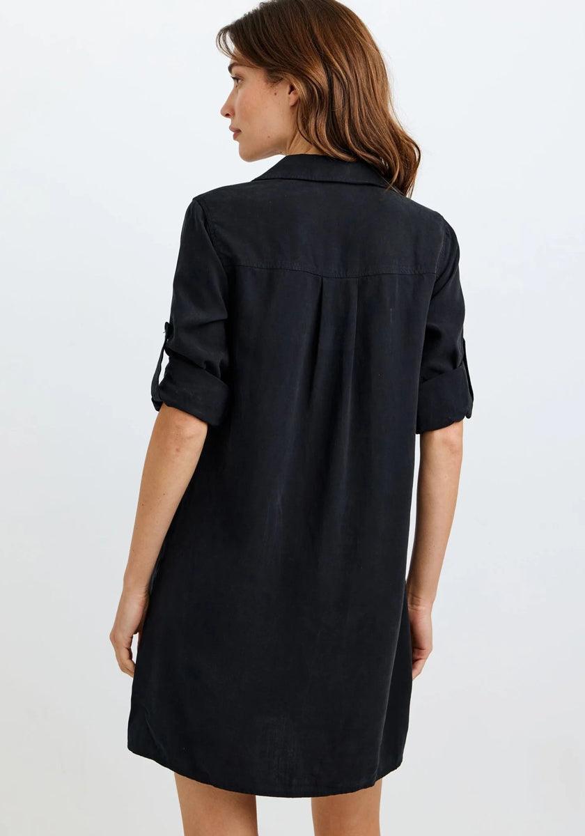 A woman with light skin and brown hair faces away, wearing a knee-length, long-sleeve Bella Dahl Lyocell dress with a loose fit and back pleat. Sleeves are rolled up and buttoned at the elbows. The background is plain white.