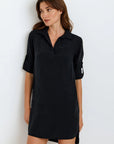 A woman with long brown hair wears a loose-fitting, black Lyocell V-neck dress with rolled-up sleeves from Bella Dahl, standing against a plain white background and gazing slightly left with a relaxed expression.