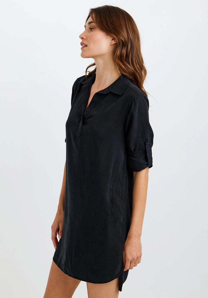 A woman with long brown hair stands sideways against a plain light background, wearing a loose-fit Bella Dahl Lyocell dress with a V-neck and rolled-up sleeves, her expression neutral.
