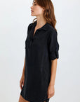 A woman with long brown hair stands sideways against a plain light background, wearing a loose-fit Bella Dahl Lyocell dress with a V-neck and rolled-up sleeves, her expression neutral.