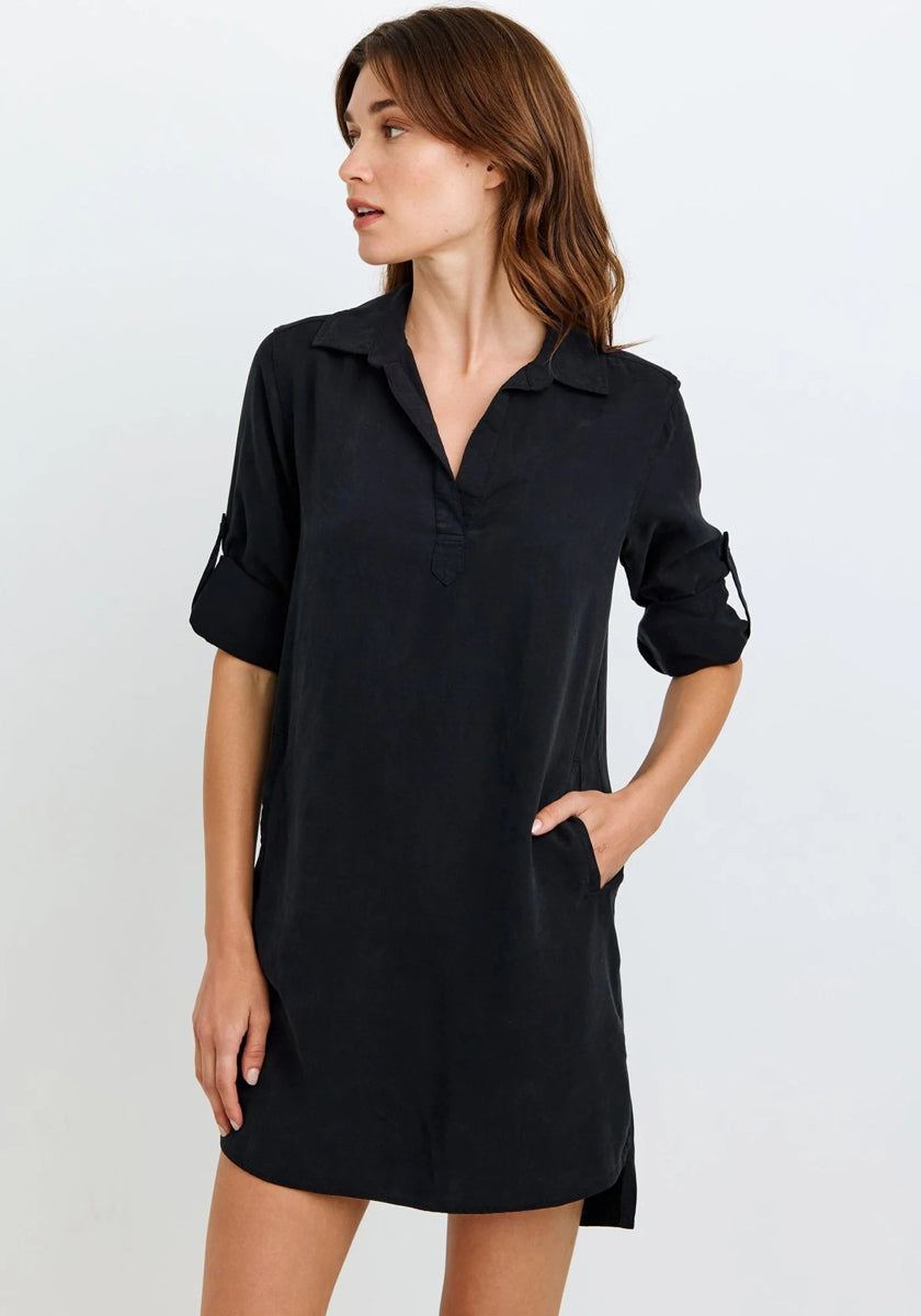 A woman with light skin and brown hair wears a loose-fitting black Lyocell V-neck dress with rolled-up sleeves from Bella Dahl. She stands sideways, one hand in the pocket, looking right against a neutral light background.
