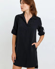 A woman with light skin and brown hair wears a loose-fitting black Lyocell V-neck dress with rolled-up sleeves from Bella Dahl. She stands sideways, one hand in the pocket, looking right against a neutral light background.