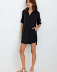 A woman stands against a white wall wearing a black V-neck Lyocell dress with rolled-up sleeves from Bella Dahl. One hand in her pocket, she pairs the look with white sandals. Her brown hair is loose and she smiles gently to the side.