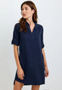 A woman with long brown hair wears a navy Bella Dahl knee-length Lyocell dress with a V-neck and rolled sleeves. She stands against a plain light background, looking left, one hand by her side, the other brushing her hip.