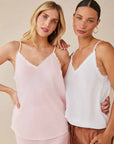 Two women smile at the camera in Bella Dahl camisoles with thin straps and frayed edges—one in pale pink with a skirt, the other in white layered over brown pants. The background is plain beige.
