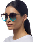 A woman with straight dark hair pulled back wears Okkias Berlino Sonnenbrille—round, oversized tortoiseshell frames with blue gradient UV 400 lenses. She wears a plain white top against a white background, facing slightly right.