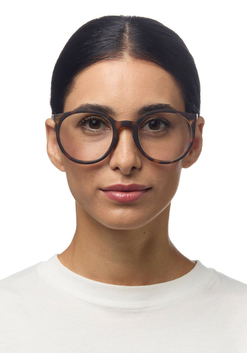 A woman with dark hair parted in the middle wears oversized Okkia reading glasses in a tortoiseshell round design and a white crew-neck shirt. Her expression is neutral, makeup is minimal, and she stands against a plain white background.