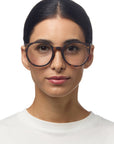 A woman with dark hair parted in the middle wears oversized Okkia reading glasses in a tortoiseshell round design and a white crew-neck shirt. Her expression is neutral, makeup is minimal, and she stands against a plain white background.