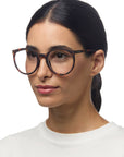 A woman with straight dark hair pulled back wears large, round Okkia reading glasses in a tortoiseshell pattern and a white top, facing slightly right with a relaxed expression against a plain white background.