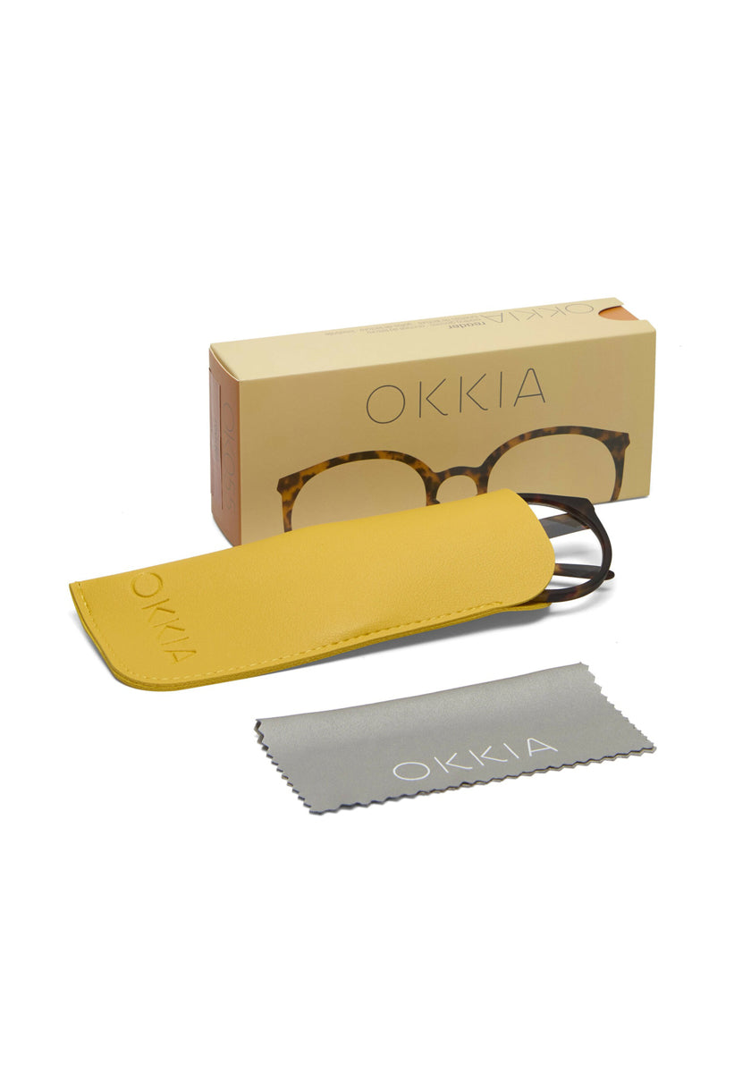 A pair of Okkia Lesebrille with Schildpattmuster frames rests partly in a yellow case. A beige Okkia box is in the background, and a gray cleaning cloth with "Okkia" in white letters is placed in the foreground.