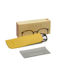 A pair of Okkia Lesebrille with Schildpattmuster frames rests partly in a yellow case. A beige Okkia box is in the background, and a gray cleaning cloth with "Okkia" in white letters is placed in the foreground.