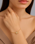 A woman with straight brown hair wears a taupe off-shoulder top by Princess Goes Hollywood and a gold-plated crab charm bracelet adorned with sparkling stones, her hand gently highlighting the jewelry against her light skin.