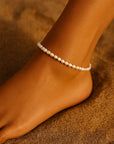 A close-up of an ankle adorned with a Princess Goes Hollywood zarte Fusskette Damen aus Süsswasserperlen. The smooth, tan skin and sunlit, golden sand evoke a warm, beachy vibe.