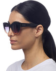 A woman with straight dark hair in a low ponytail wears black Okkia cat-eye sunglasses with UV400 protection. She has clear skin and a white top, shown in profile against a plain white background.