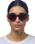 A woman with straight dark hair parted in the middle wears Okkia Cat-Eye-Stil Sonnenbrille with tortoiseshell frame and UV 400-Schutz. She has a neutral expression and is dressed in a plain white crew-neck top against a white background.
