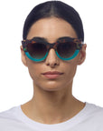 A woman with straight, dark hair in a white shirt wears Okkia’s bold Cat-Eye-Sonnenbrille featuring tortoiseshell frames and turquoise bottoms. She faces forward neutrally; these retro Sonnenbrillen offer UV 400 protection.