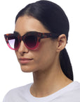 A woman with straight dark hair tied back wears oversized Okkia Cat-Eye sunglasses with a red and tortoiseshell frame. She has a neutral expression and is dressed in a plain white crew-neck shirt against a white background.