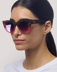 Eine Person mit glattem, nach hinten gezogenem dunklem Haar trägt eine Okkia Cat-Eye-Sonnenbrille in Schildpatt und Rosa mit UV-400-Schutz, gepaart mit einem weißen Oberteil, das von den Schultern aufwärts im Dreiviertelprofil vor einem schlichten hellen Hintergrund gezeigt wird.