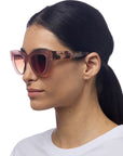 A woman with long dark hair pulled back wears pink gradient cat-eye sunglasses from Okkia and a white crew-neck shirt. She faces left, revealing the Okkia logo on the arm. The look exudes retro elegance against a plain white background.