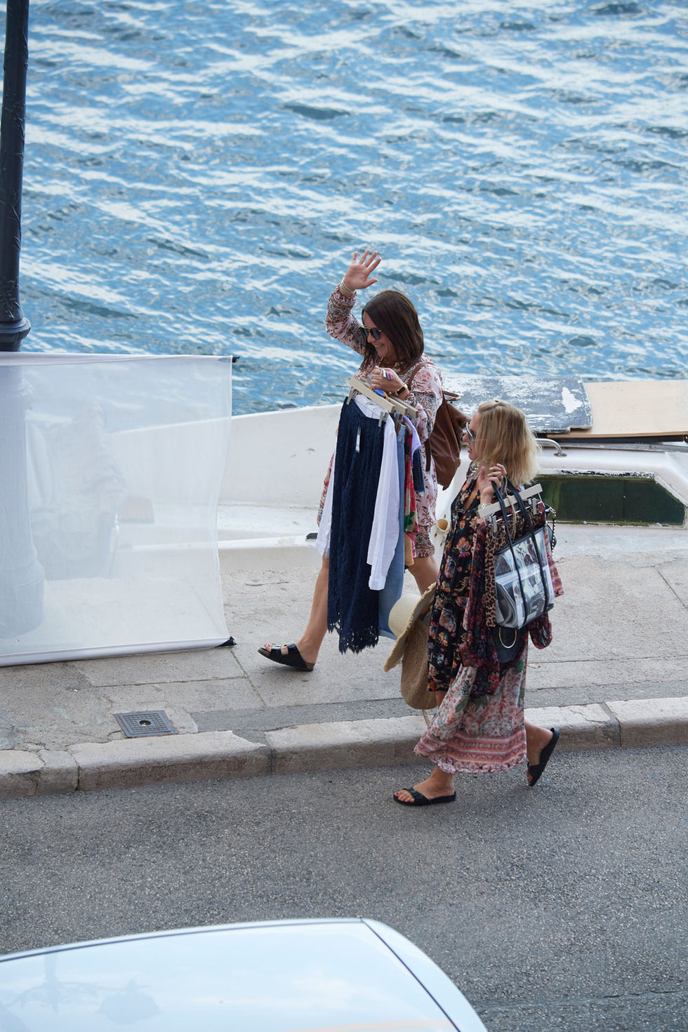 Two women in patterned dresses walk along a waterfront sidewalk. One woman waves while holding several hanging clothes; the other carries a bag and has sunglasses. The blue water and part of a car are visible in the background.