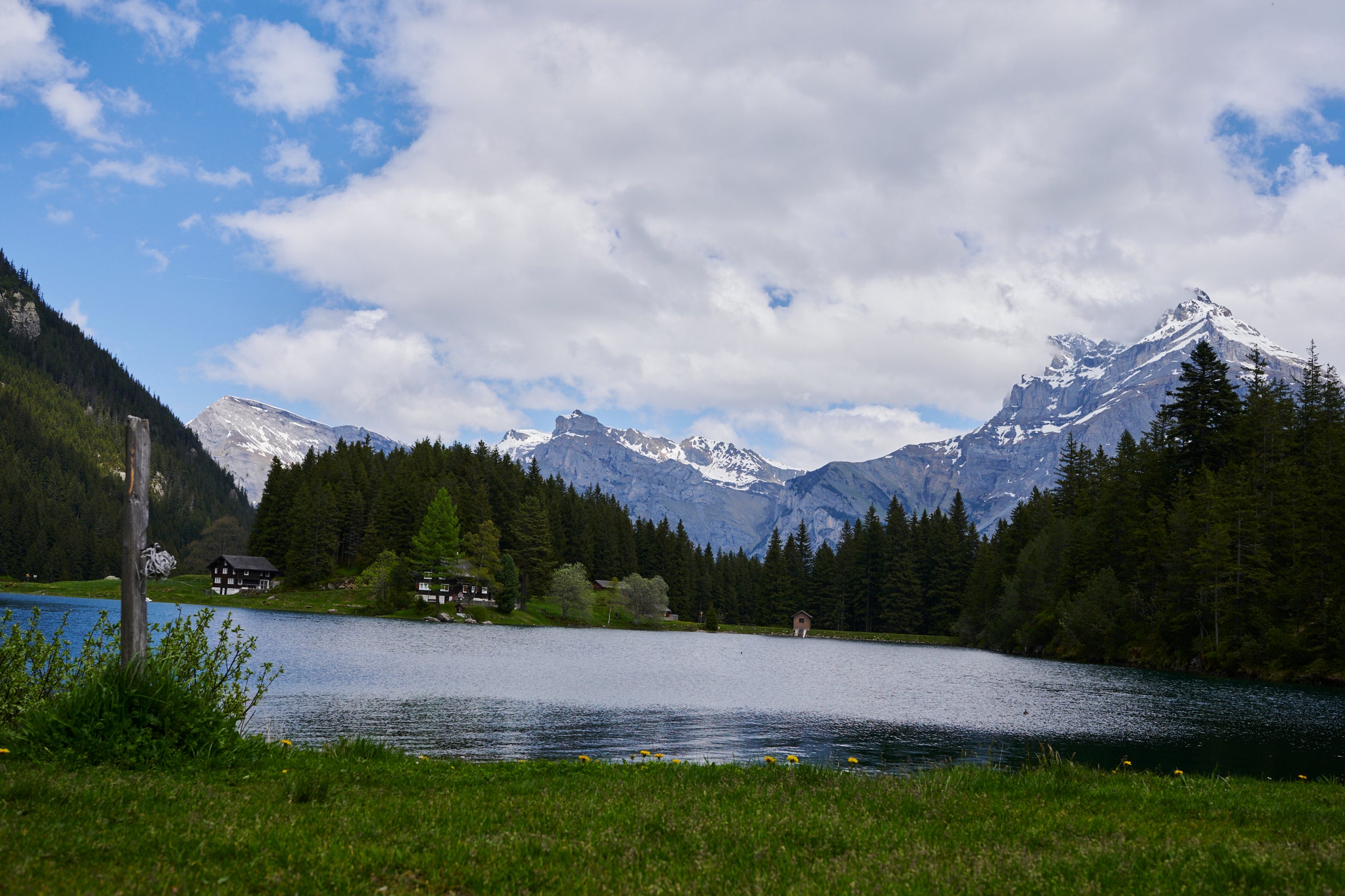 A tranquil lake reflects surrounding pine trees and snow-capped mountains under a partly cloudy sky. Alpine cabins are nestled among the trees, and green grass lines the lakeshore in the foreground. The scene feels peaceful and natural.