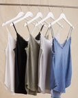 Five Bella Dahl sleeveless tops—denimoptik, breathable Lyocell, and more—hang on white hangers in white, black, olive green, light gray, and denim blue against a beige fabric background.