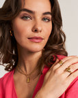 A woman with wavy brown hair wears a bright pink top, gold hoop earrings, two layered 18K Vergoldung necklaces, and the Princess Goes Hollywood Vergoldeter Ring in Tropfenform. She looks confidently at the camera with her hand on her shoulder.