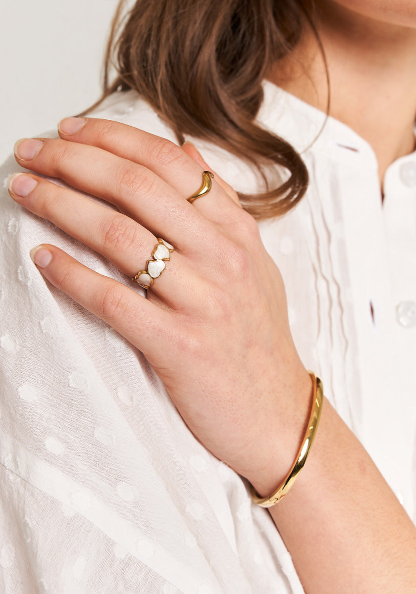 A woman’s hand wearing Princess Goes Hollywood’s “Ring mit Herzen in Perlmutt-Optik” and a gold bracelet rests on her chest. She has wavy brown hair and wears a textured white blouse.
