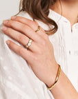 A woman’s hand wearing Princess Goes Hollywood’s “Ring mit Herzen in Perlmutt-Optik” and a gold bracelet rests on her chest. She has wavy brown hair and wears a textured white blouse.